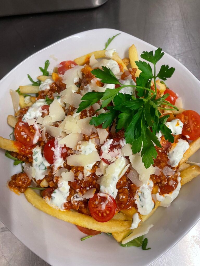 Eine Portion kanadische Poutine auf einem weißen Teller. Goldbraune Pommes frites sind mit einer Hackfleisch-Tomaten-Sauce, weißen Saucenklecksen, halbierten Kirschtomaten und gehobeltem Hartkäse garniert. In der Mitte befindet sich ein frischer Zweig glatte Petersilie. Am unteren Bildrand steht der Text ‚Kanadisches Poutine‘