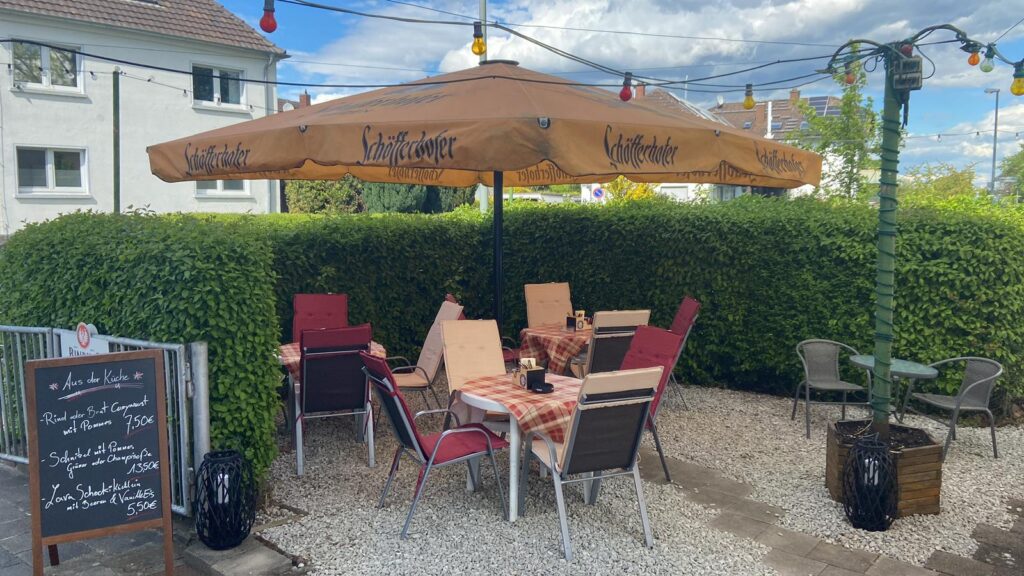 Ein gemütlicher Außenbereich eines Restaurants auf Kiesboden. Ein großer, beigefarbener Sonnenschirm überspannt mehrere Tische mit rot-weiß karierten Decken und Stühlen mit roten Polstern. Im Vordergrund steht eine hölzerne Kreidetafel mit handgeschriebener Speisekarte. Der Bereich ist von einer dichten grünen Hecke und einer Lichterkette umgeben.