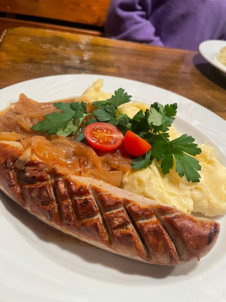 Eine angerichtete Bratwurst mit rautenförmigem Grillmuster auf einem weißen Teller. Daneben liegt cremiger Kartoffelstampf, getoppt mit glänzenden Schmorzwiebeln, einer halbierten Kirschtomate und einem frischen Zweig glatte Petersilie. Am unteren Bildrand steht der Text ‚Bratwurst mit Kartoffelstampf und Schmorzwiebeln‘