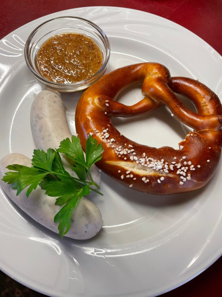 Ein klassisches bayerisches Frühstück auf einem weißen Teller: Zwei Weißwürste, garniert mit einem Zweig glatter Petersilie, daneben eine ofenfrische Laugenbrezel mit grobem Salz. Dazu wird in einem kleinen Glasschälchen süßer Senf serviert. Am unteren Bildrand steht der Text ‚Weißwurst, Süßer Senf und Brezel‘