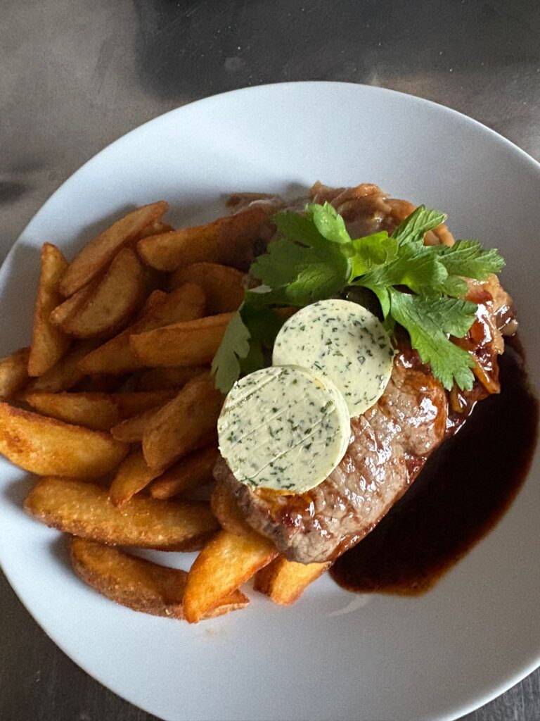 Ein angerichtetes Rumpsteak auf einem weißen Teller, serviert mit goldbraunen Kartoffelecken. Das Fleisch ist mit einer dunklen Sauce unterlegt und mit zwei runden Scheiben Kräuterbutter sowie einem frischen Zweig glatte Petersilie garniert. Am unteren Bildrand steht der Text ‚Rumpsteak mit Kartoffelecken‘